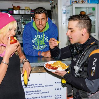 Zwei Taucher stehen nebeneinander am Imbiss, die eine isst einen Apfel, der andere eine Currywurst.