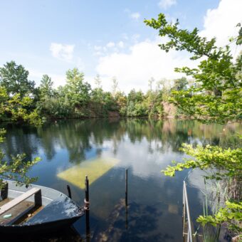 Blick über den von Bäumen und Felswänden gesäumten Steinbruchsee Pinckvos, vorne ein Ruderboot, im klaren Wasser schimmert eine Unterwasserplattform.