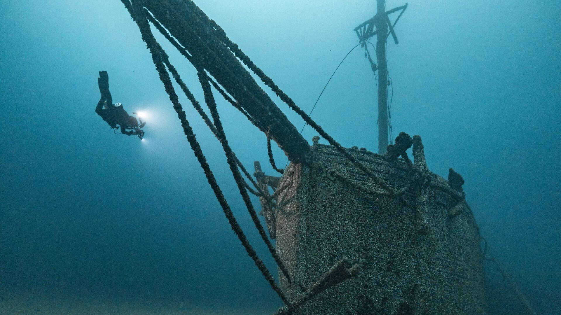 Taucher mit Lampe schwebt neben dem bewachsenen Bug eines Segelschiffwracks, von dem ein langes Rundholz schräg nach oben ragt.