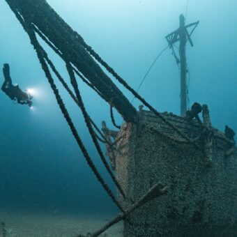 Taucher mit Lampe schwebt neben dem bewachsenen Bug eines Segelschiffwracks, von dem ein langes Rundholz schräg nach oben ragt.