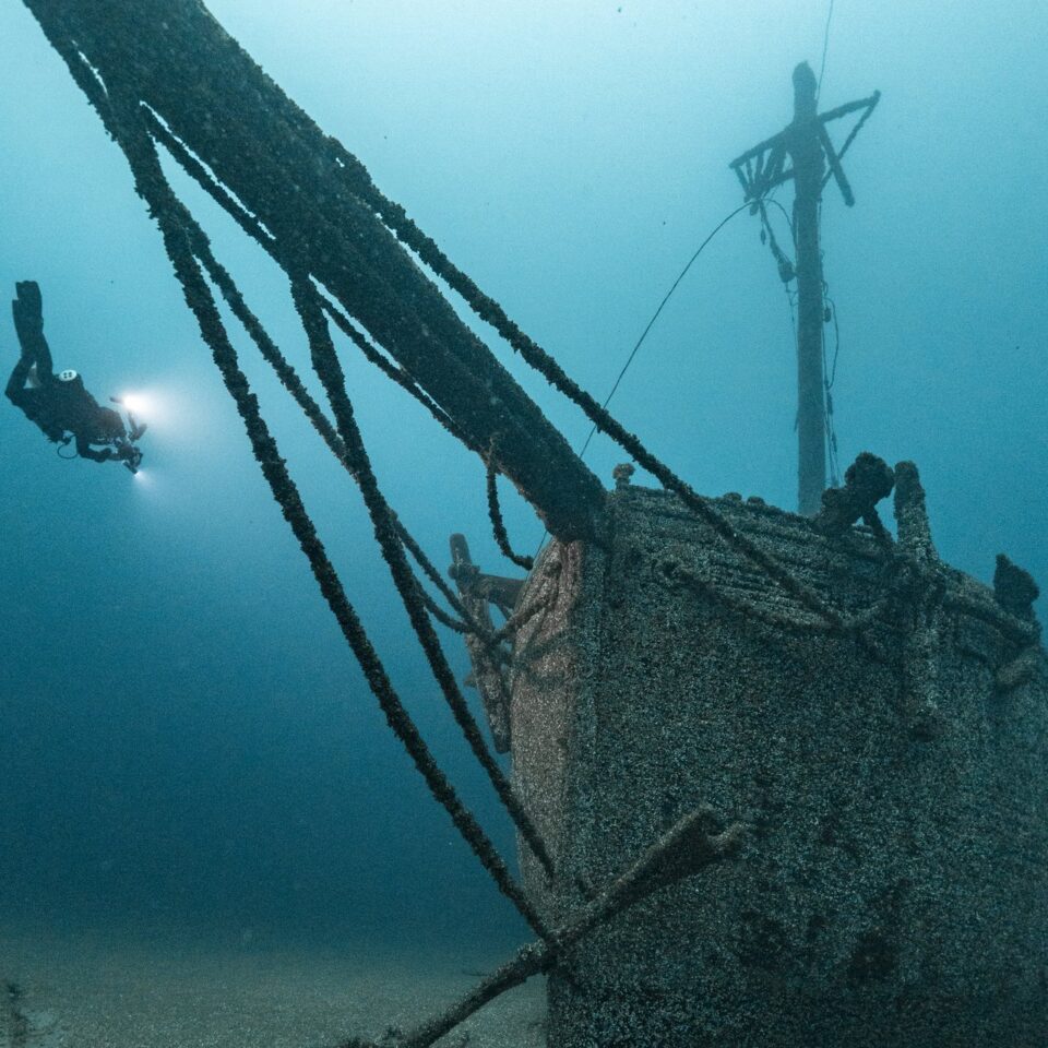 Taucher mit Lampe schwebt neben dem bewachsenen Bug eines Segelschiffwracks, von dem ein langes Rundholz schräg nach oben ragt.