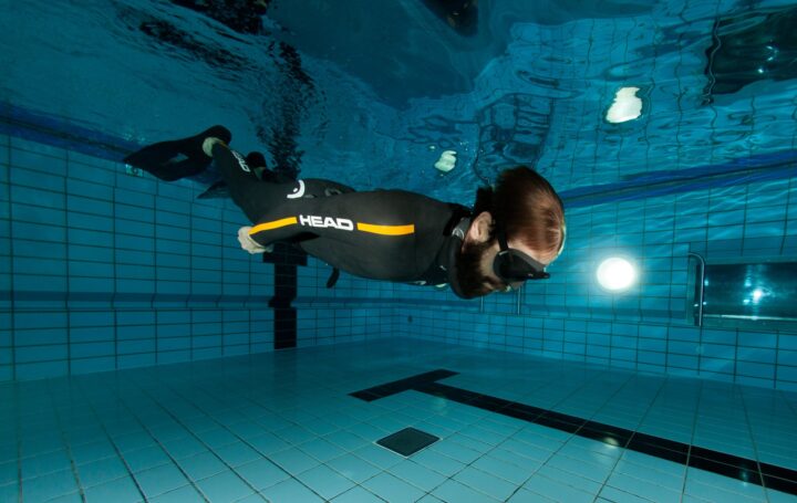 Apnoetaucher im Neoprenanzug schwebt mit angelegten Armen waagerecht im Schwimmbecken, im Hintergrund eine Unterwasserlampe