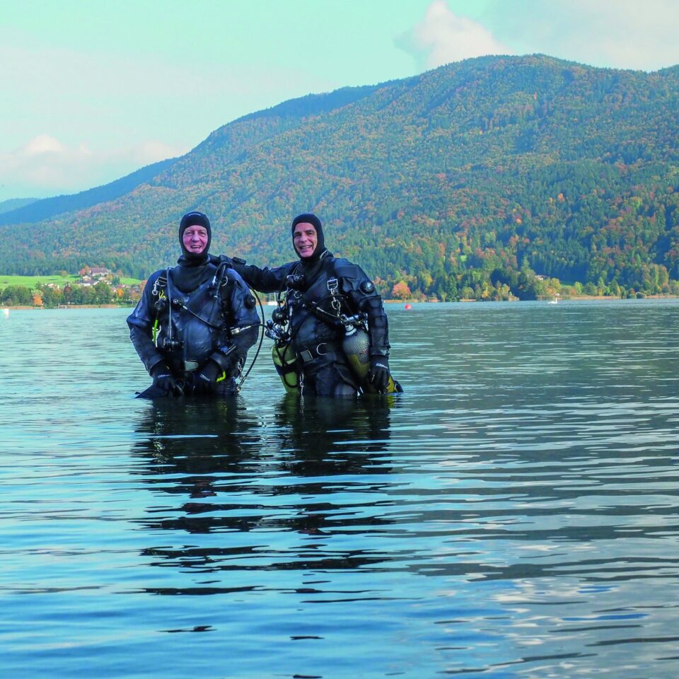 Zwei Taucher in Camaro-Trockentauchanzügen mit Tauchgeräten stehen bauchtief im Mondsee, dahinter ein bewaldeter Berg