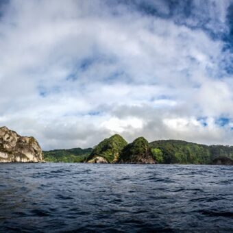 Panorama von Cocos Island mit steilen, grün bewachsenen Felsen über dem Meer unter Wolkenhimmel