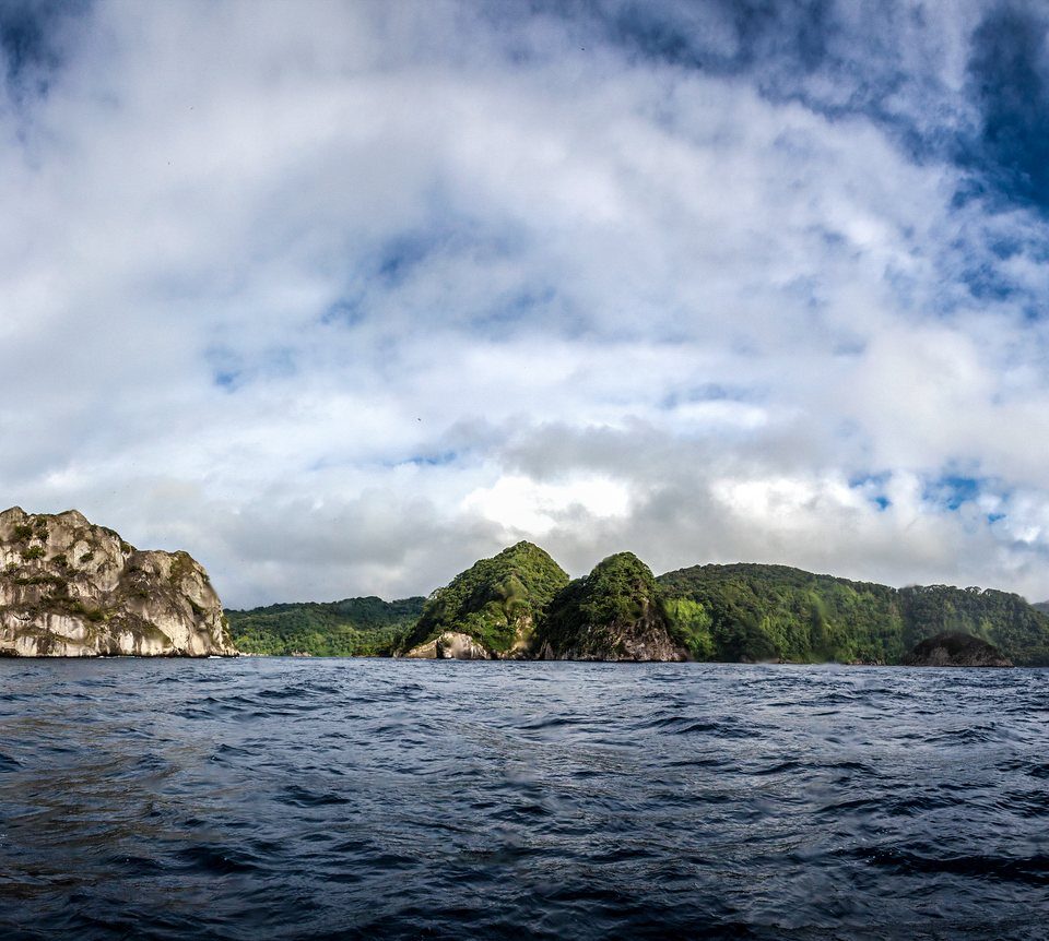 Panorama von Cocos Island mit steilen, grün bewachsenen Felsen über dem Meer unter Wolkenhimmel