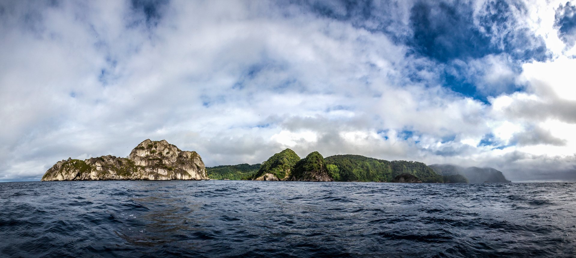 Panorama von Cocos Island mit steilen, grün bewachsenen Felsen über dem Meer unter Wolkenhimmel